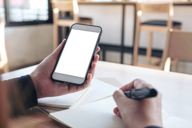 istock Mockup image of hands holding white mobile phone with blank white screen while writing on notebook in office 1059444356