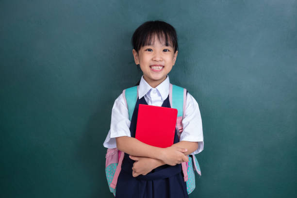 azjatycka chińska dziewczynka w mundurku niosąca torbę szkolną i książkę przeciwko zielonej tablicy - male education college student nerd zdjęcia i obrazy z banku zdjęć
