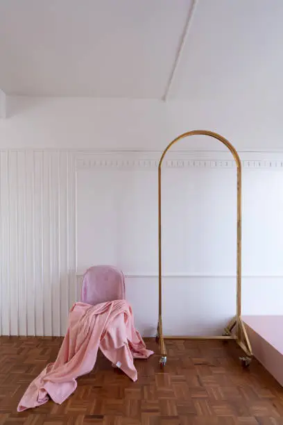 Composition of oldrose blanket setting on mid century modern chair in baby pink color setting in white wooden stripe and classic moulding wall and gold clothes hanger.