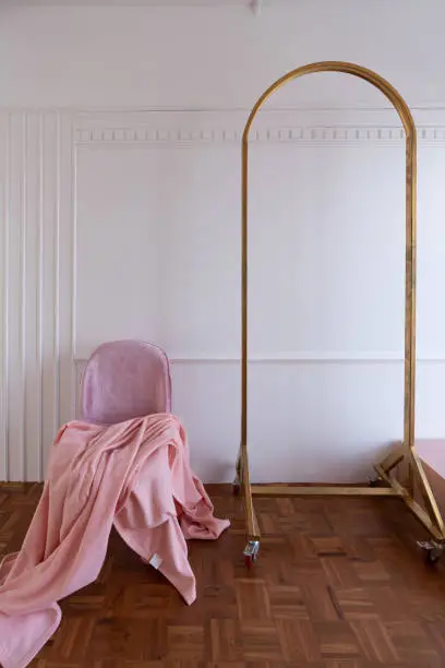 Composition of oldrose blanket setting on mid century modern chair in baby pink color setting in white wooden stripe and classic moulding wall and gold clothes hanger.