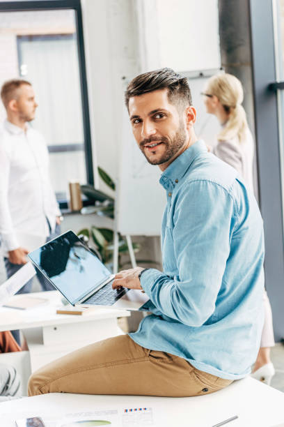 istock handsome young businessman using laptop and smiling at camera in office 1059551402
