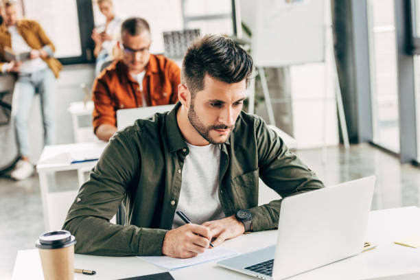 istock thoughtful young man working with laptop and notepad at open space office with colleagues on background 1059563308