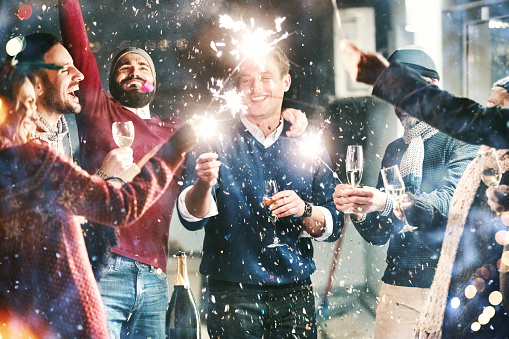 Closeup of group of mixed age people having New year's party on a balcony. They are having champagne, lighting up some fire sparklers and popping confetti. There are three women and four men enjoying this party. One of the women is obscured.

High ISO shot.