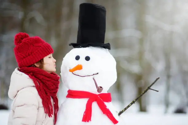 Photo of Adorable little girl building a snowman in beautiful winter park. Cute child playing in a snow.