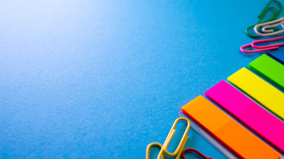 Multicolored paper clips and bookmarks on blue background. View from above with copy space