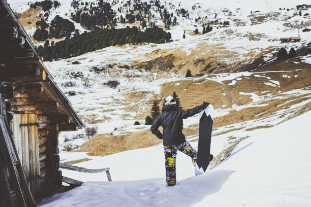 istock Snowboarder wearing black jacket, mask with snowboard close authentic wooden house. Horizontal. 1059599680