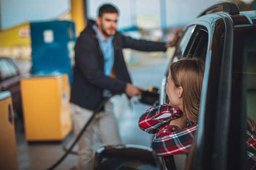 Man and woman, man pouring gasoline in his car at the gas station, woman is sitting in a car.