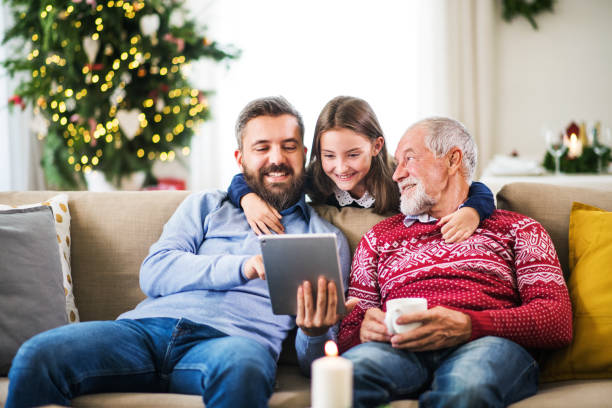 Small girl and her father and grandfather sitting on a sofa at Christmas, using tablet. A happy small girl and her father and grandfather sitting on a sofa at Christmas time, using tablet. multi generation family christmas stock pictures, royalty-free photos & images