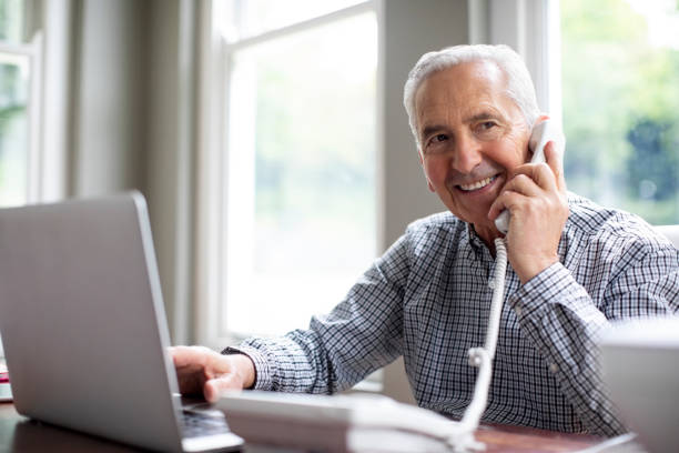 Smiling man talking on phone while using laptop Smiling senior man talking through telephone while looking away at desk. Happy retired male is sitting with laptop against window in domestic room. He is in formalwear at home. landline phone stock pictures, royalty-free photos & images