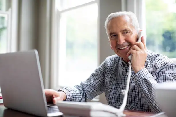 Smiling senior man talking through telephone while looking away at desk. Happy retired male is sitting with laptop against window in domestic room. He is in formalwear at home.