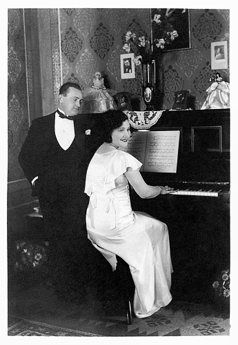 Vintage black and white photo of the thirties featuring a woman playing piano while the man standing is looking at her.