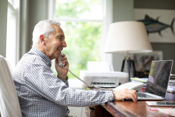 Smiling man talking on phone while using laptop Smiling senior man talking through telephone while using laptop at desk. Side view of retired male is sitting in domestic room. He is in formalwear at home. landline phone stock pictures, royalty-free photos & images