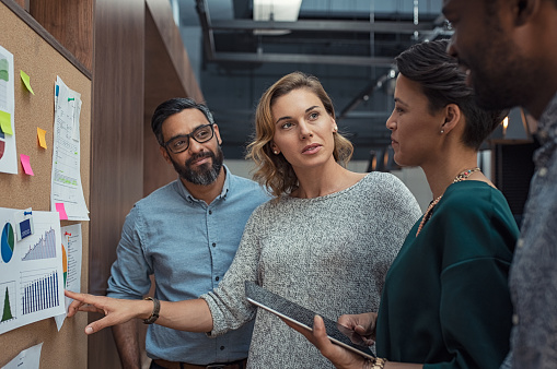 Multiethnic business people discussing charts and graphs showing the results of their successful teamwork. Mature business woman showing presentation to casual colleagues in modern office during night meeting. Group of creatives analyzing market trends.