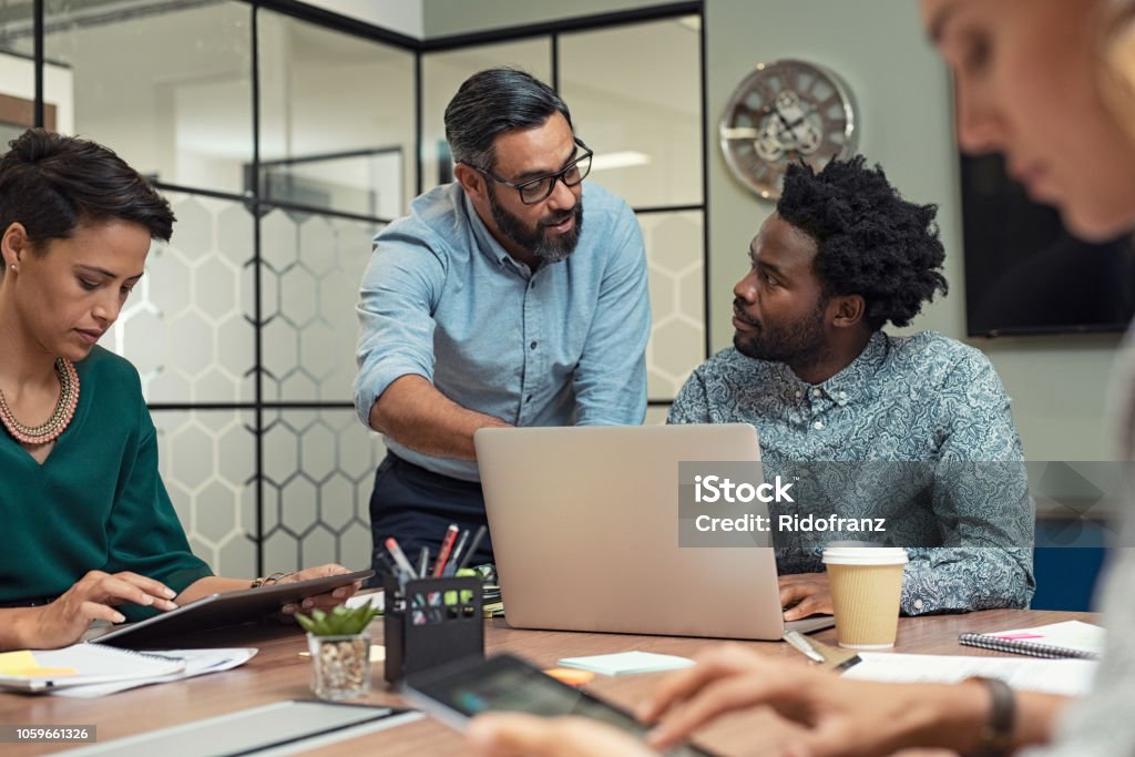 Business people working in a meeting room Latin mature manager discussing with his colleague online data in front of laptop. Serious businessman wearing eyeglasses training employees in job. Creative multiethnic team working together with boss in a meeting room. Education Training Class Stock Photo