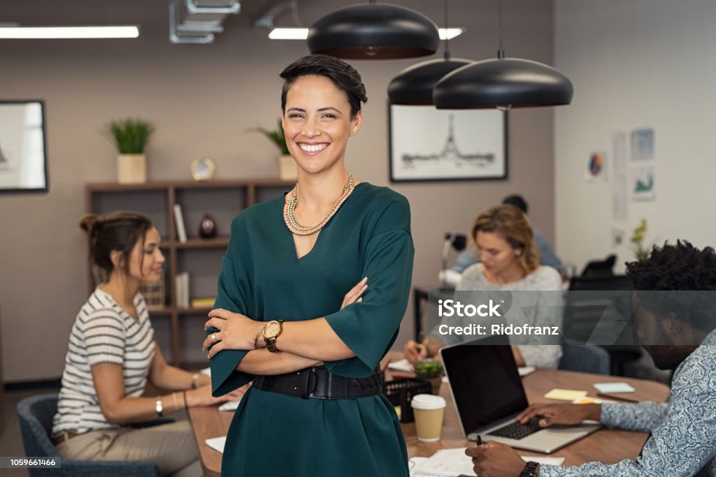 Successful young business woman Portrait of successful business woman standing with her colleagues working in background at office. Portrait of cheerful fashion girl in green dress standing with folded arms and looking at camera. Beautiful businesswoman feeling proud and smiling. Leadership Stock Photo