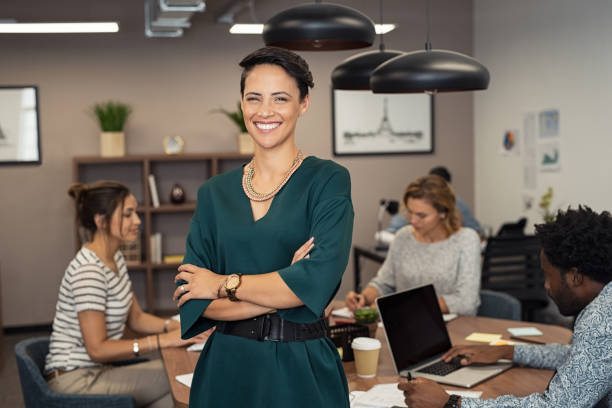 Successful young business woman Portrait of successful business woman standing with her colleagues working in background at office. Portrait of cheerful fashion girl in green dress standing with folded arms and looking at camera. Beautiful businesswoman feeling proud and smiling. business casual fashion stock pictures, royalty-free photos & images