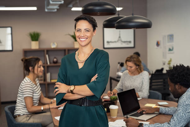 Successful young business woman Portrait of successful business woman standing with her colleagues working in background at office. Portrait of cheerful fashion girl in green dress standing with folded arms and looking at camera. Beautiful businesswoman feeling proud and smiling. woman arms folded stock pictures, royalty-free photos & images