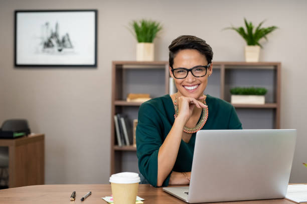 Successful business woman smiling Portrait of happy casual businesswoman sitting at her desk in office. Cheerful latin woman working on laptop and looking at camera. Young fashionable girl wearing eyeglasses sitting at creative agency. secretary stock pictures, royalty-free photos & images