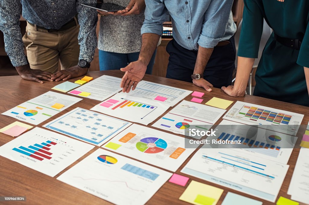 Business casual team working on graphs Financial and business documents on table with multiethnic hands working on it. Latin business manager with colleagues working on new startup project. Closeup business man and businesswoman hands understanding pie and bar graphs during meeting. Data Stock Photo
