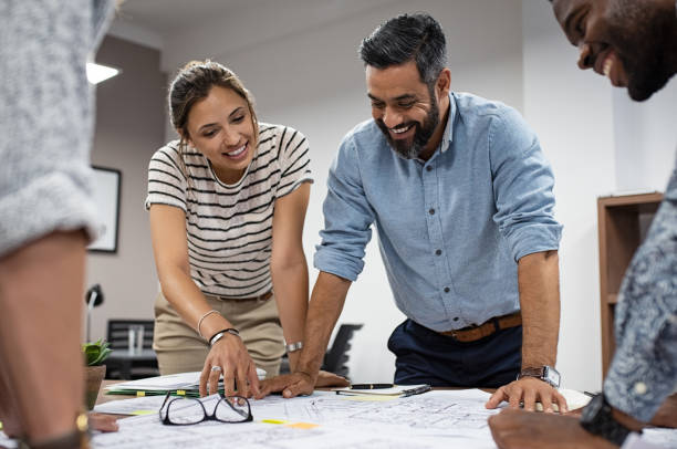 Group of architects working together Team of multiethnic architects working on construction plans in meeting room. Cheerful engineers discussing on project in office. Group of mature businessman and young woman standing around table working on blueprint. architect stock pictures, royalty-free photos & images