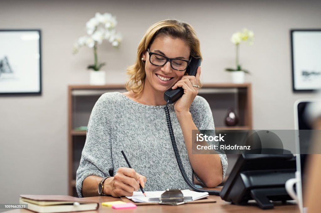 Happy mature woman talking on phone Beautiful mature woman talking on phone at creative office. Happy smiling businesswoman answering telephone at office desk. Casual business woman sitting at her desk making telephone call and taking notes on notebook. Telephone Stock Photo