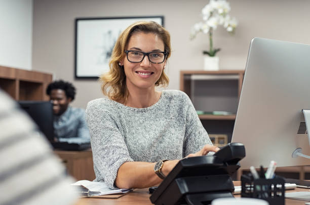 Mature casual woman working on computer Mature beautiful woman working at creative office and looking at camera. Portrait of receptionist working on computer in a coworking space. Successful businesswoman with eyeglasses sitting at help desk and smiling. secretary stock pictures, royalty-free photos & images