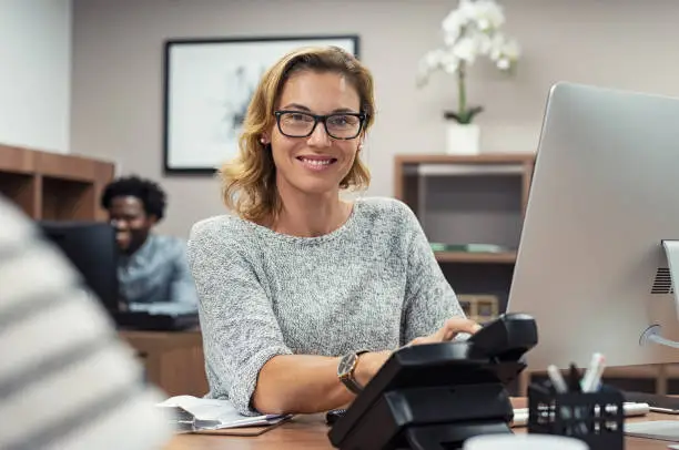 Mature beautiful woman working at creative office and looking at camera. Portrait of receptionist working on computer in a coworking space. Successful businesswoman with eyeglasses sitting at help desk and smiling.
