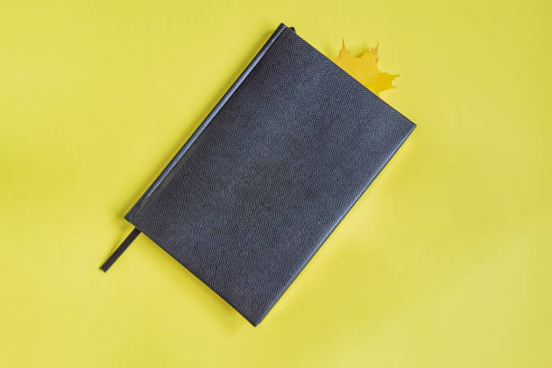 istock False reptile leather notebook of black color with maple leaf as bookmark on yellow background. Minimal flat lay 1059696528