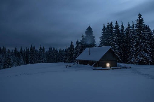 Wooden cabin under stars. Lights shines through the window from inside of the house. Night landscape in winter. Smoke goes from the chimney.