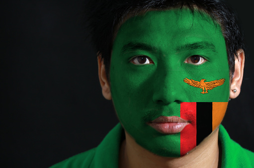 Portrait of a man with the flag of the Zambia painted on his face on black background, A green field with an orange colored eagle in flight over a rectangular block of red black and orange.
