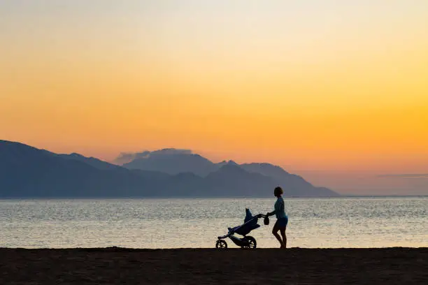 Mother with baby stroller enjoying motherhood at sunset landscape. Walking or jogging woman with pram on a beach at sunset. Beautiful inspirational mountains landscape.