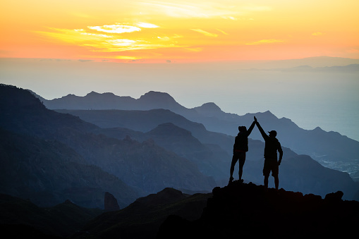 Teamwork couple helping hand trust help, silhouette success in mountains. Team of climbers man and woman. Hikers celebrate with hands up, help each other on top of mountain, climbing together, beautiful sunset landscape.