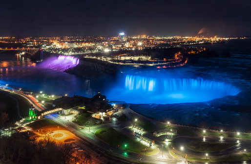 A high angle view of Niagara falls at night, with Horseshoe Falls lit up in blue, and the American Falls in purple.