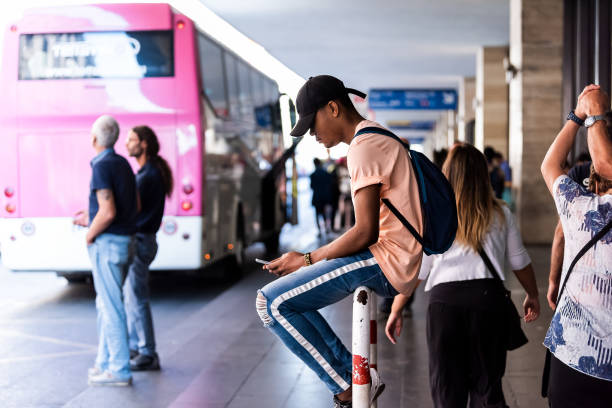młody człowiek siedzący z telefonem przy wejściu do dworca kolejowego termini, letni dzień, ludzie turyści chodzący na pokład czekając na autobus terravision na lotnisko - ciampino airport zdjęcia i obrazy z banku zdjęć