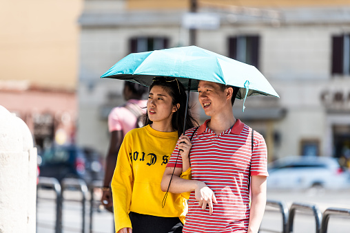 Rome, Italy - September 4, 2018: Historic city with young asian couple under umbrella shade walking by Colosseum, sunny summer day, people romantic on street