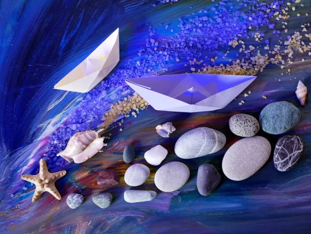 istock Decorative composition installation - smooth sea stones, sea salt, shells, starfish and paper boats on a blue background 1059717374