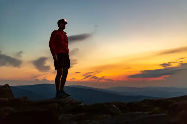 Trail runner with head lamp. Man celebrating sunset on mountain top. Looking at inspiring view. Hiker or climber reached mountain peak, enjoy inspiring landscape on rocky path Karkonosze, Poland