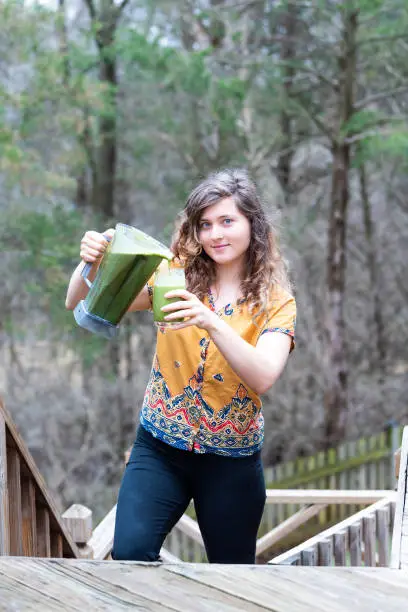 Young woman standing outside, outdoors, holding plastic blender container, pouring green vegetable, kale smoothie into drinking glass on wooden deck