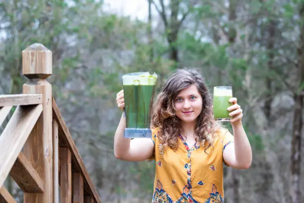 Young woman outside, outdoors, holding plastic blender container for smoothie with green vegetables in drinking glass on wooden home, house, deck