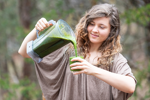 Young woman standing outside, outdoors, holding plastic blender container, pouring green vegetable, kale smoothie into drinking glass