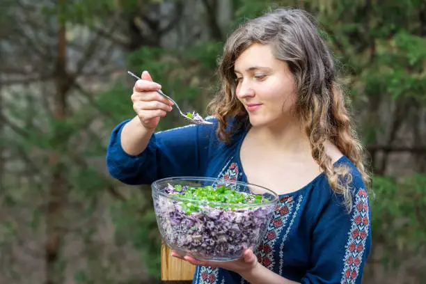 Young woman outside, outdoors, holding glass bowl of homemade red, purple cabbage salad dish with green onions, scallions with spoon in hand, eating