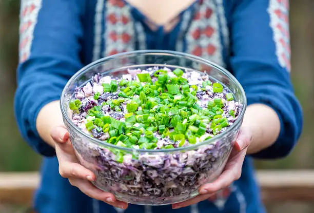 Closeup of young woman showing, offering outside, outdoors, holding glass bowl of homemade red, purple cabbage salad dish with green onions, scallions