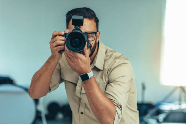 Portrait of a photographer taking photo standing in a studio Photographer standing in position to take a photograph. Man looking into a digital camera for taking  a photograph. photographer stock pictures, royalty-free photos & images