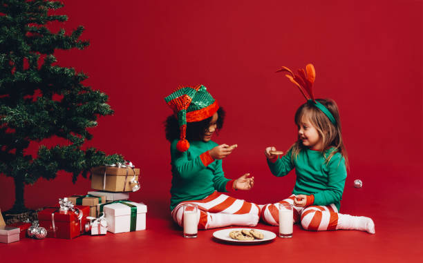 Little girls sitting beside a christmas tree enjoying cookies and milk. Two happy kids dressed in christmas theme clothes sitting on floor beside a christmas tree and gift boxes.