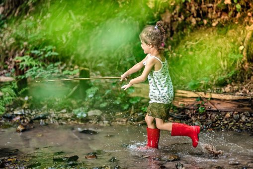 Little smiling girl in red rubber boots running across stream in the forest holding wood stick.