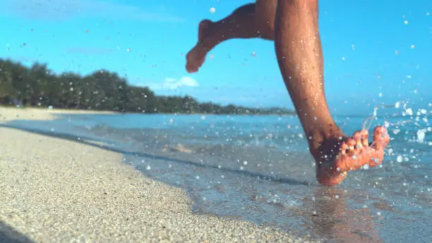 COPY SPACE, LOW ANGLE, DOF: Unrecognizable male tourist jogs on the empty tropical beach and splashes seawater. Active young man on holiday runs barefoot in the wet sand on the coast of exotic island.