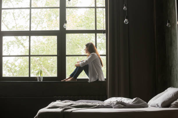 istock Lonely sad young woman sitting on windowsill at home 1059754818