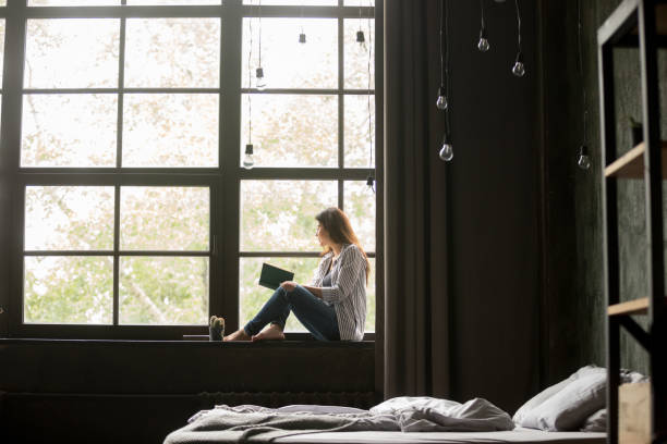 istock Woman with book sitting on window sill at home 1059754834