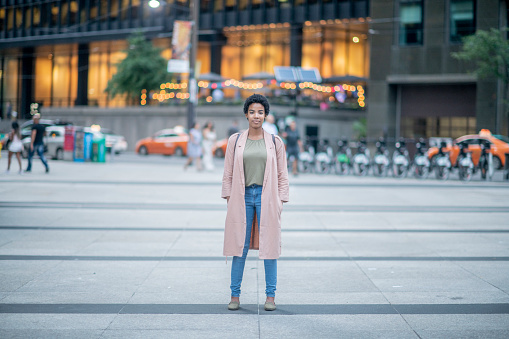 A woman of African descent is standing and posing in street of a large city. She is wearing stylish clothing.