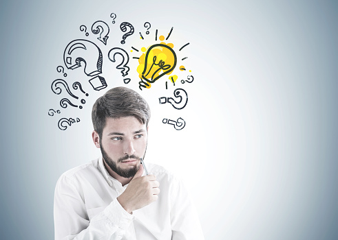 Troubled young bearded caucasian businessman in white shirt sitting with pen near his face and thinking looking sideways. A gray wall background with question marks and a yellow light bulb. Mock up